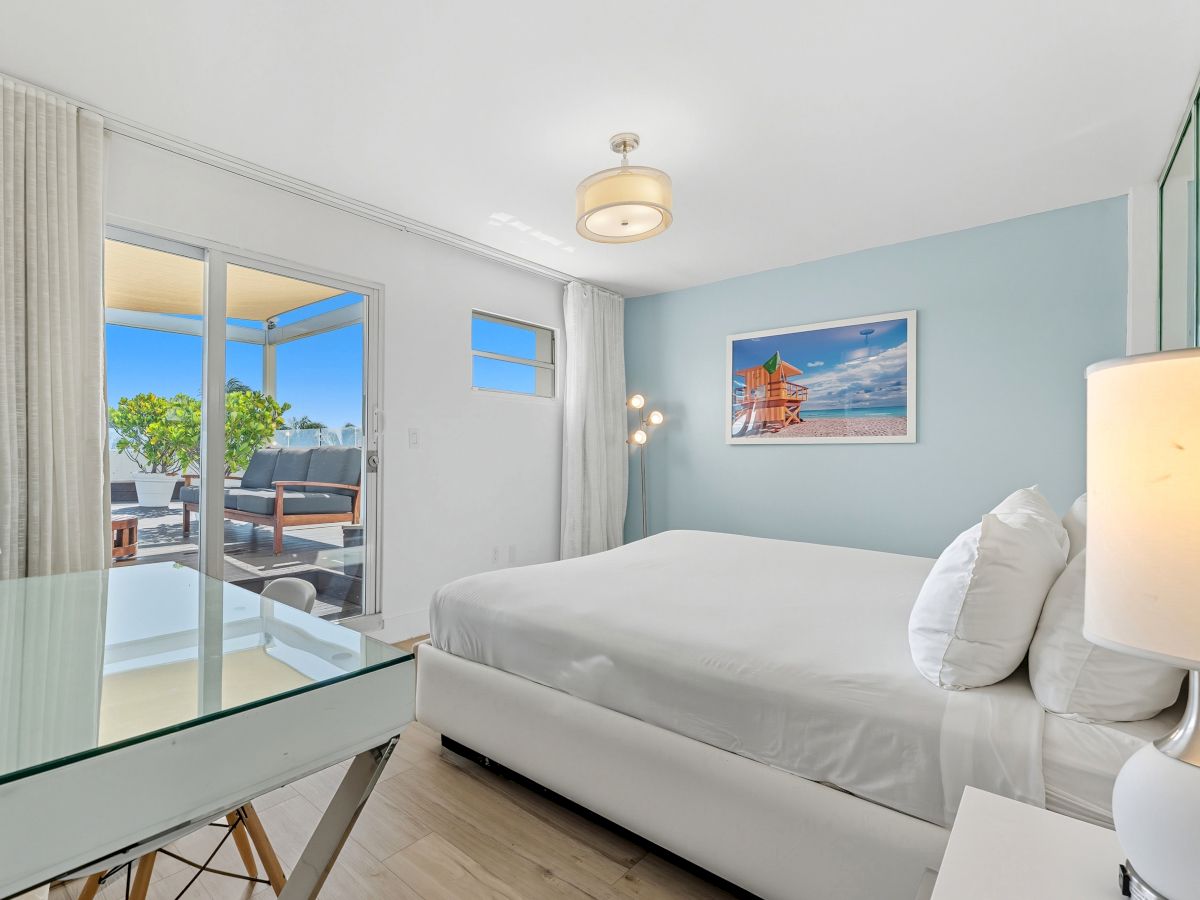 A modern bedroom with a bed, desk, and lamp, featuring a balcony with outdoor seating and a framed picture on the wall.