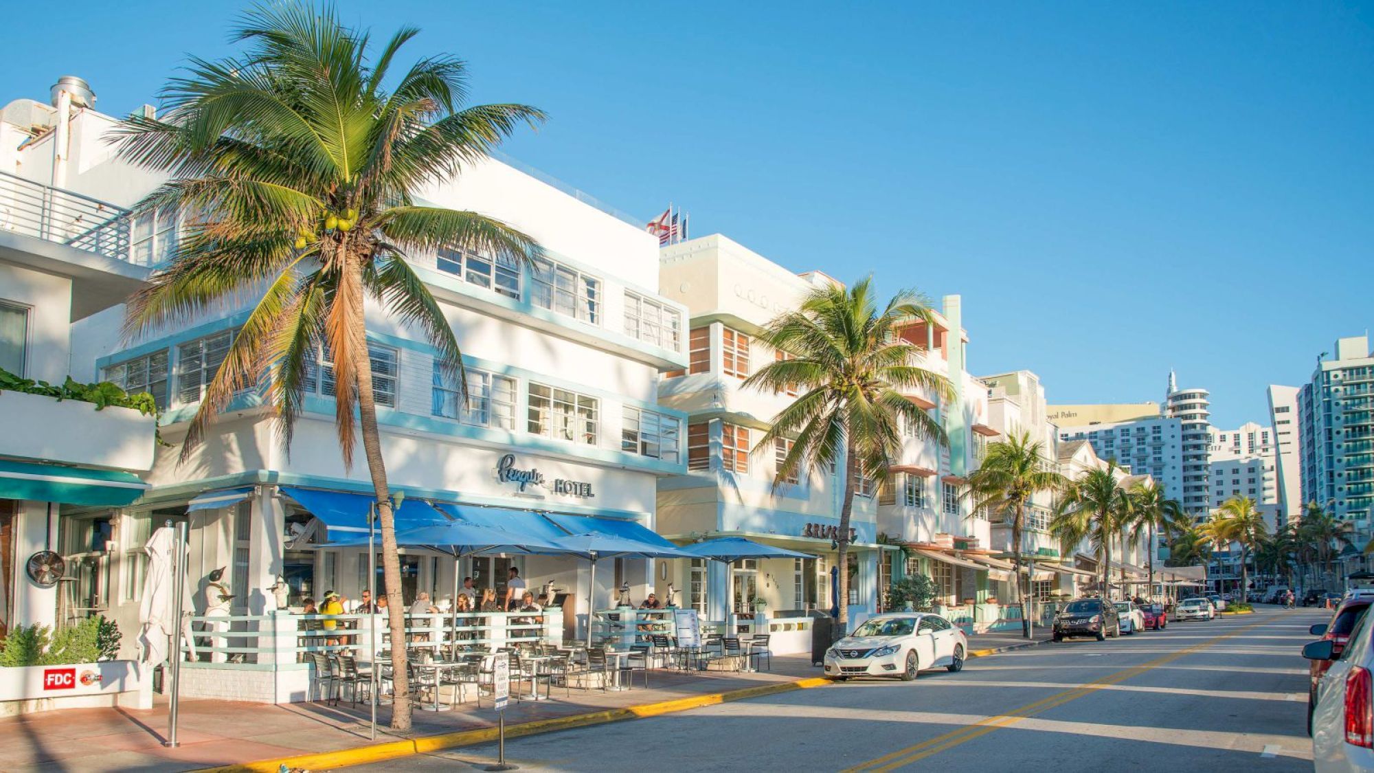 A sunny street with palm trees, pastel-colored buildings, shops, restaurants, and parked cars, capturing a vibrant urban scene on a clear day.