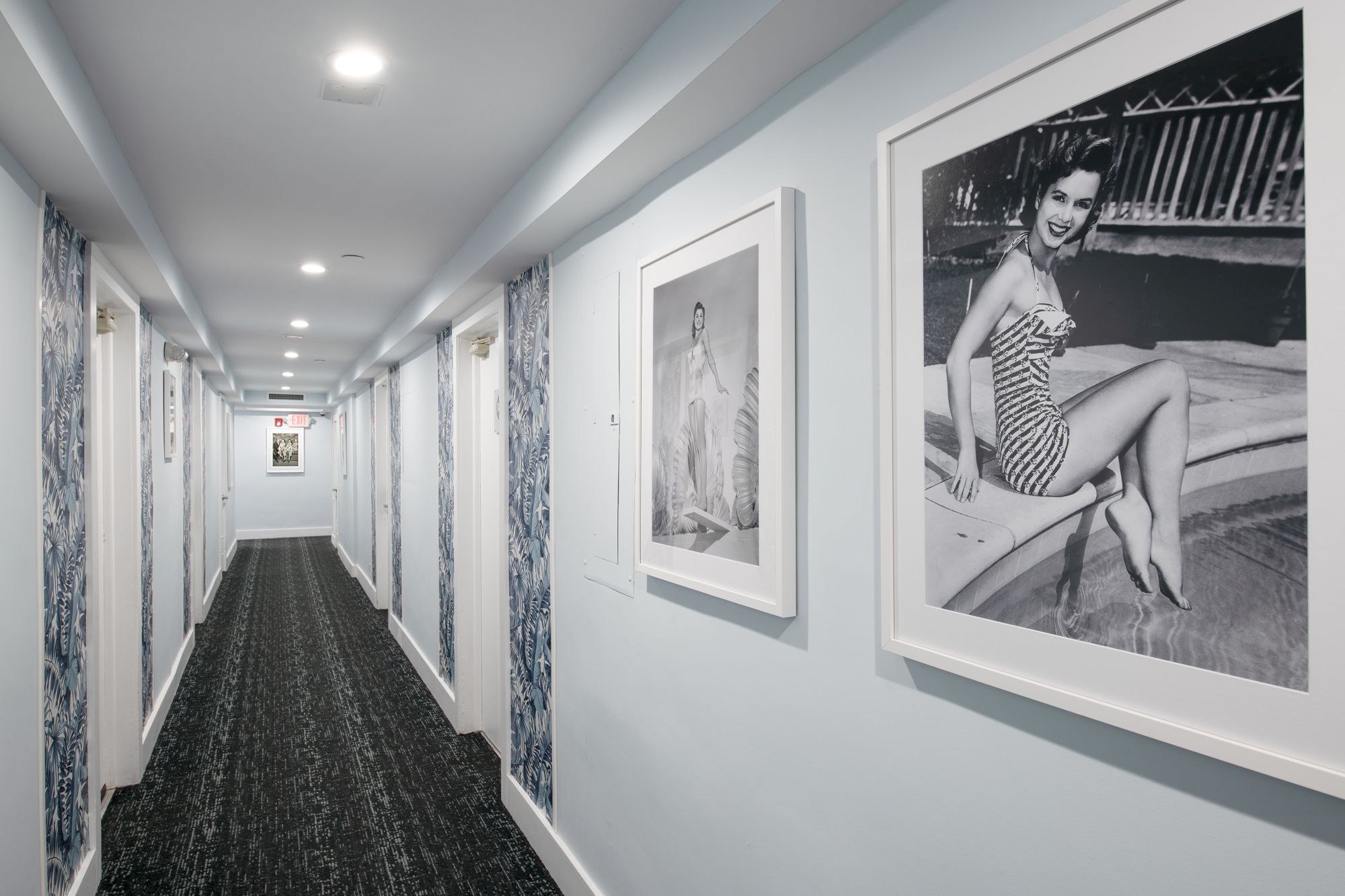 A hotel hallway with framed black-and-white vintage photographs on light blue walls, a patterned carpet, and recessed ceiling lights.