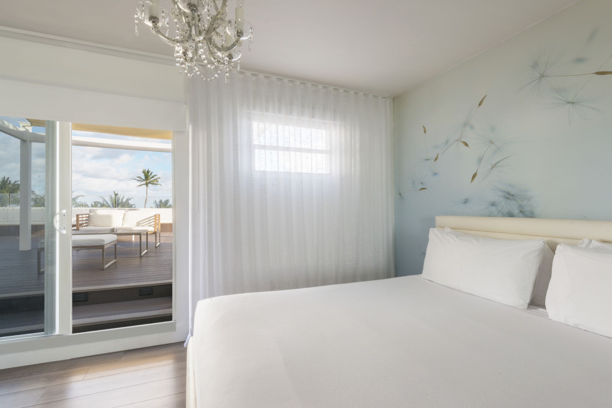 A bright bedroom with a white bed, chandelier, sheer curtains, and wall mural, leading to a balcony with outdoor seating.