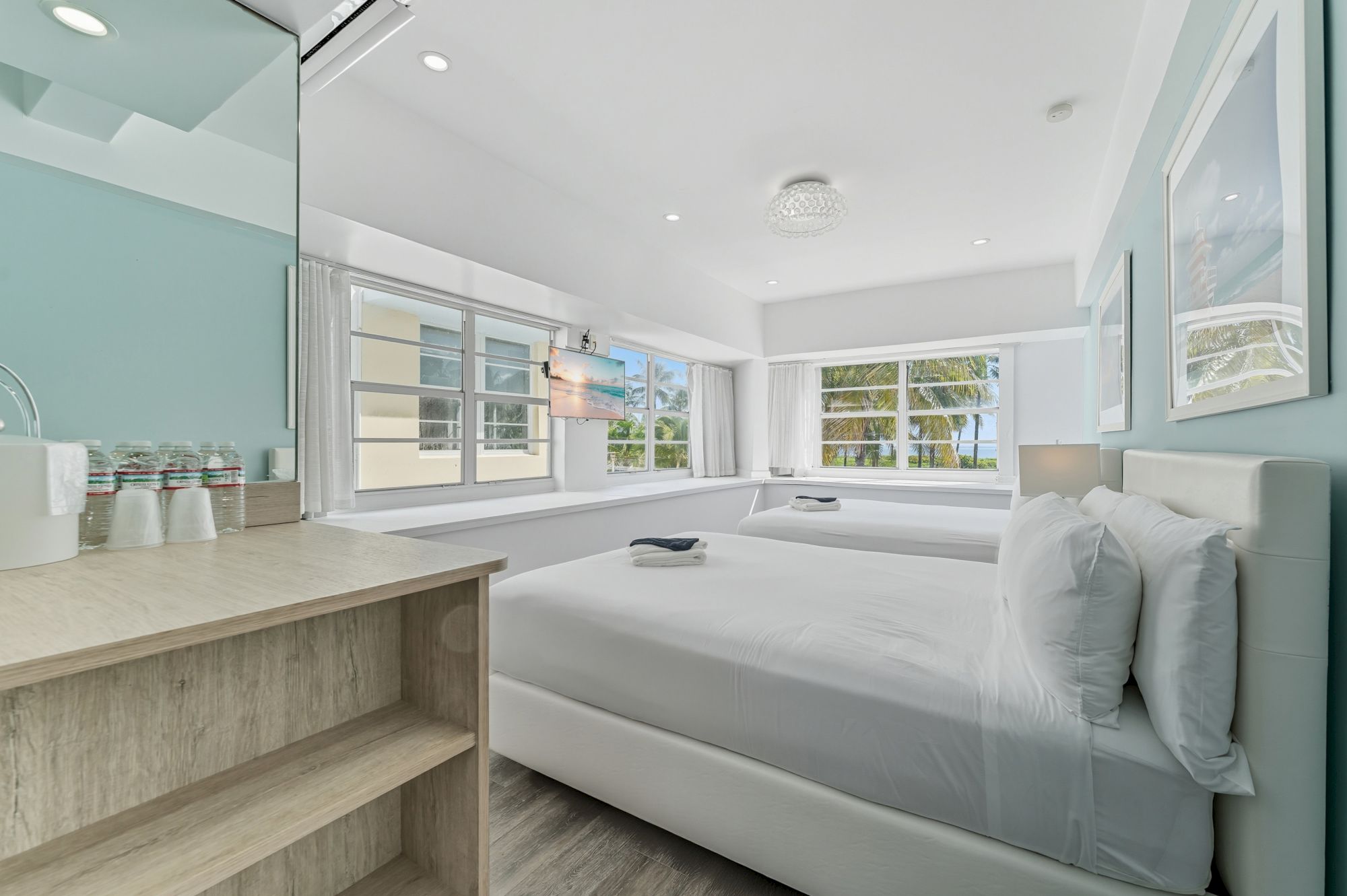 A bright, modern bedroom with two beds, a dresser with toiletries, and large windows letting in natural light complete the scene.