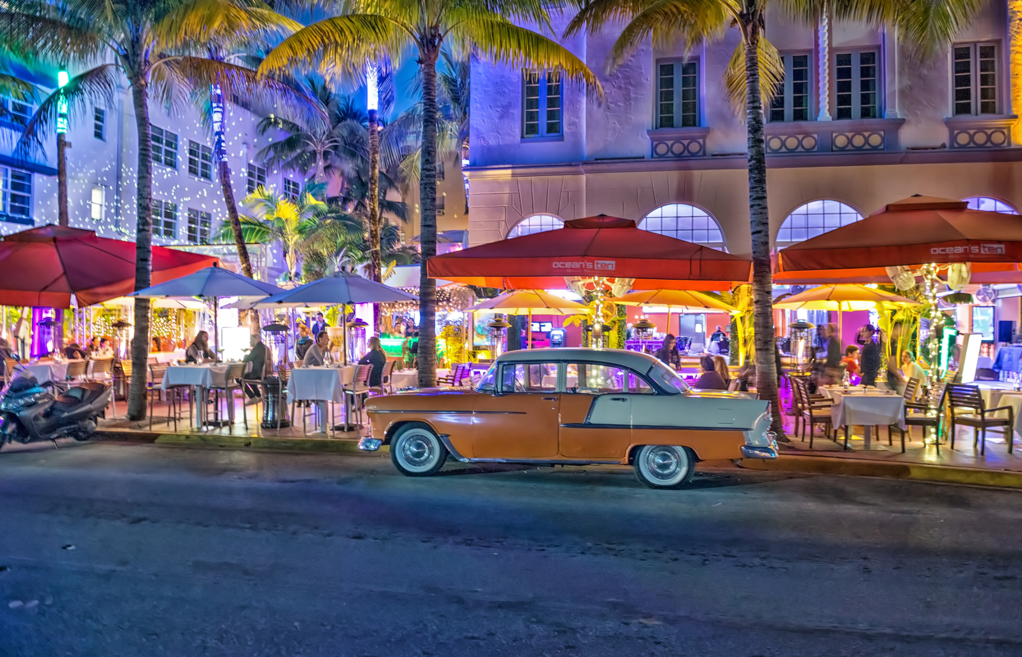 A vibrant street scene with a classic car, outdoor dining areas with red umbrellas, illuminated palm trees, and colorful lights at night in an urban setting.