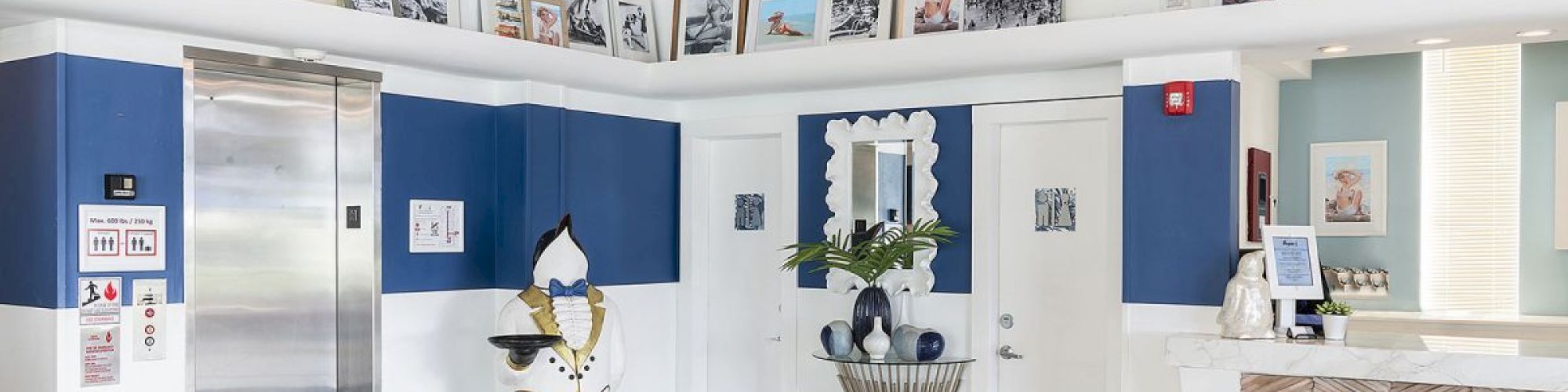 The image shows a stylish lobby with a blend of blue and white decor, featuring an elevator, framed pictures, a sculpted penguin, and a reception desk.