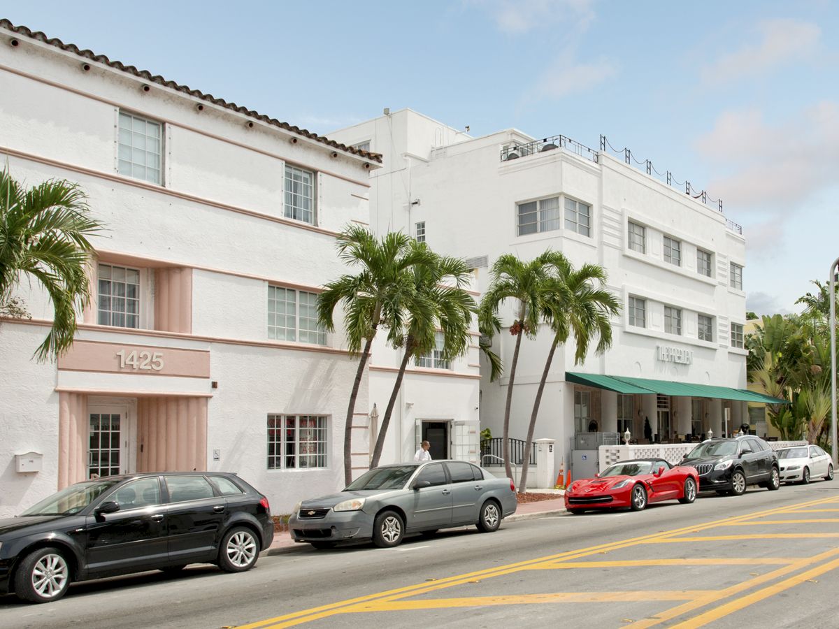 A street with parked cars, palm trees, and white buildings; one with visible signage. The image shows a vibrant, sunny day.