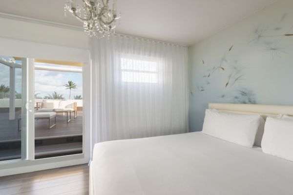 A bright, minimalist bedroom with a large bed, white linens, a chandelier, and a balcony with a seating area outside the glass door and window.