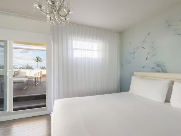 A bright, minimalist bedroom with a large bed, white linens, a chandelier, and a balcony with a seating area outside the glass door and window.