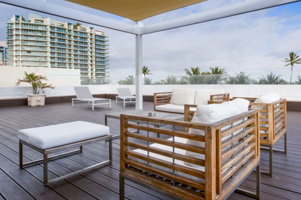 An outdoor patio with modern wooden furniture, white cushions, lounge chairs, and potted plants overlooking nearby buildings and palm trees.