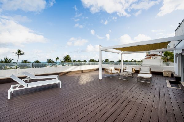 A spacious outdoor deck with lounge chairs, seating arrangements under a pergola, and a scenic view of palm trees and a blue sky with clouds.