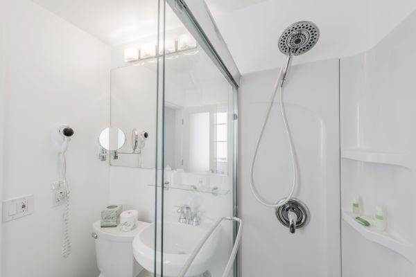 A modern bathroom with a shower, sink, mirror, and toilet. It has white walls and fixtures, a wall-mounted hair dryer, and shelf for toiletries.