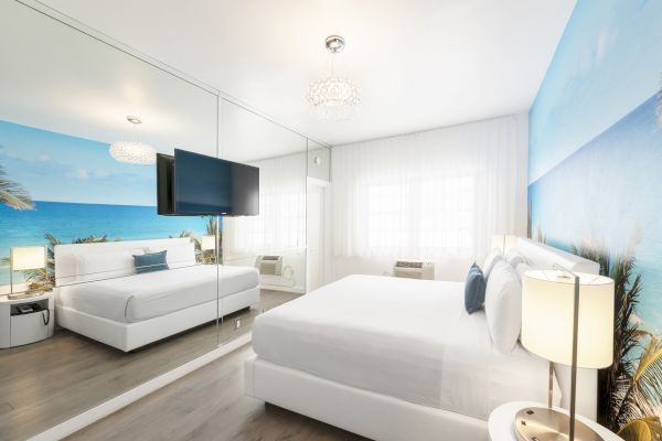 A modern bedroom has a large mirror, a wall mural of a beach, a flat-screen TV, and large windows with white curtains.