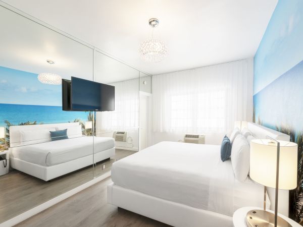 A modern bedroom has a large mirror, a wall mural of a beach, a flat-screen TV, and large windows with white curtains.