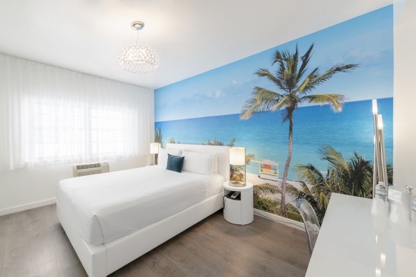 A modern bedroom with a white bed, a chandelier, and a wall mural depicting a tropical beach scene with palm trees and clear blue water ending the sentence.