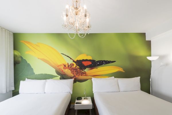 The image shows a hotel room with two beds, a chandelier, a floor lamp, and a large mural of a butterfly on a yellow flower behind the beds.