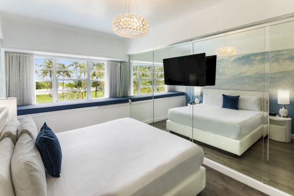 The image shows a modern bedroom with large windows, a mirrored wall, a TV, two beds, and neutral decor with blue accents and scenic outdoor views.