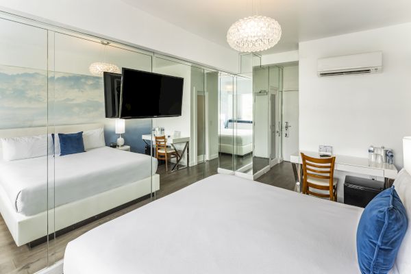 A modern hotel room featuring two beds with blue pillows, a large TV, mirrored walls, a desk with a chair, and an air conditioning unit.