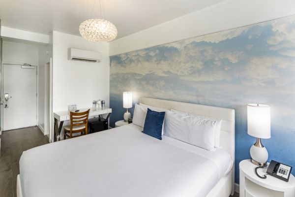 The image shows a modern hotel room with a large bed, wall art of clouds, a desk, lamps, air conditioning unit, and a door to what looks like a bathroom.