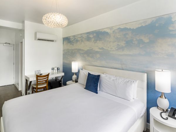 The image shows a modern hotel room with a large bed, wall art of clouds, a desk, lamps, air conditioning unit, and a door to what looks like a bathroom.