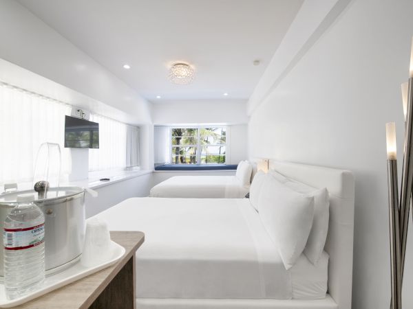 A modern hotel room features two beds, a mounted TV, a bottle of water with two glasses on a tray, and a window view with natural light.