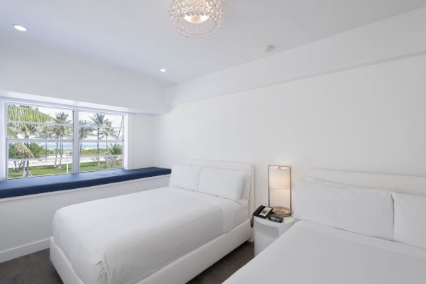 A bright, clean hotel room with two white beds, a window seat, a bedside table with a lamp, and a large window overlooking palm trees and water.