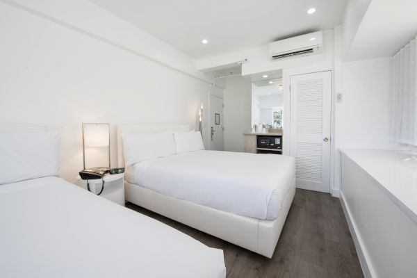 A modern, white-themed hotel room with two beds, a nightstand with a lamp, an air conditioner, a small kitchen area, and minimal decor.