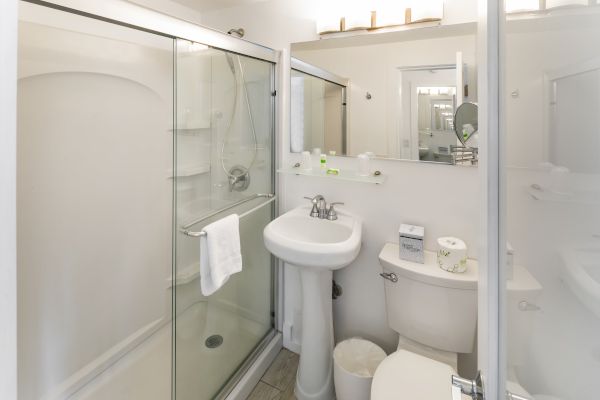 A compact bathroom with a glass shower enclosure, pedestal sink, mirror, toilet, and various toiletries neatly arranged on a shelf.