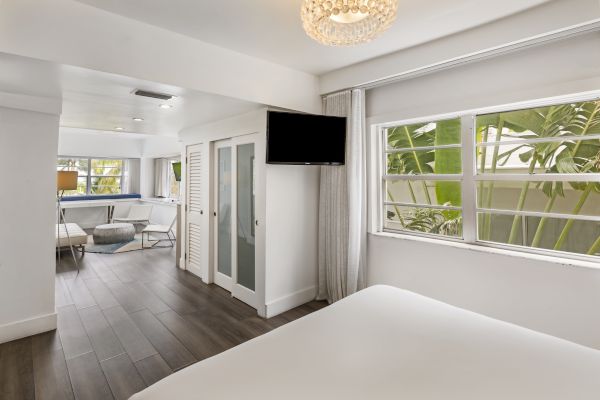 A modern, bright bedroom with a large window, flat-screen TV, and adjacent seating area, featuring stylish, minimalistic décor and wooden flooring.