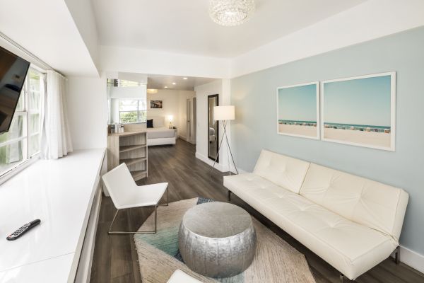 A modern living room with a white sofa, chair, round silver ottoman, and TV, featuring beach-themed artwork on the wall and a lamp in the corner.