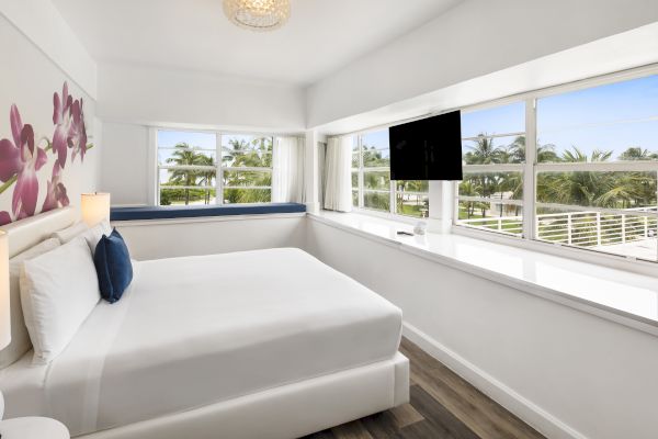 A modern bedroom with a white bed, a floral wall design, large windows with a view, a TV, and a blue bench by the window at the far end.