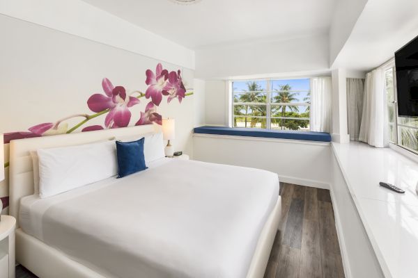 A bright, modern bedroom with a large bed, floral wall art, window seat, and a scenic view of palm trees through the window, with a TV on the wall.