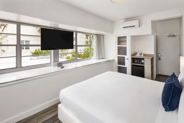 A modern bedroom with large windows, a wall-mounted TV, a bed with white linens and navy pillows, a kitchenette, and a door.