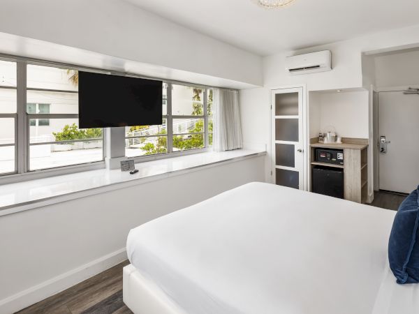 A modern bedroom with large windows, a wall-mounted TV, a bed with white linens and navy pillows, a kitchenette, and a door.