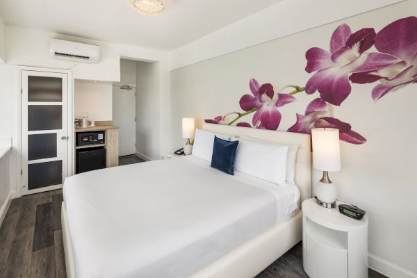 A modern hotel room with a bed, purple floral wall art, two bedside lamps, an air conditioner, a small kitchenette, and wooden flooring.