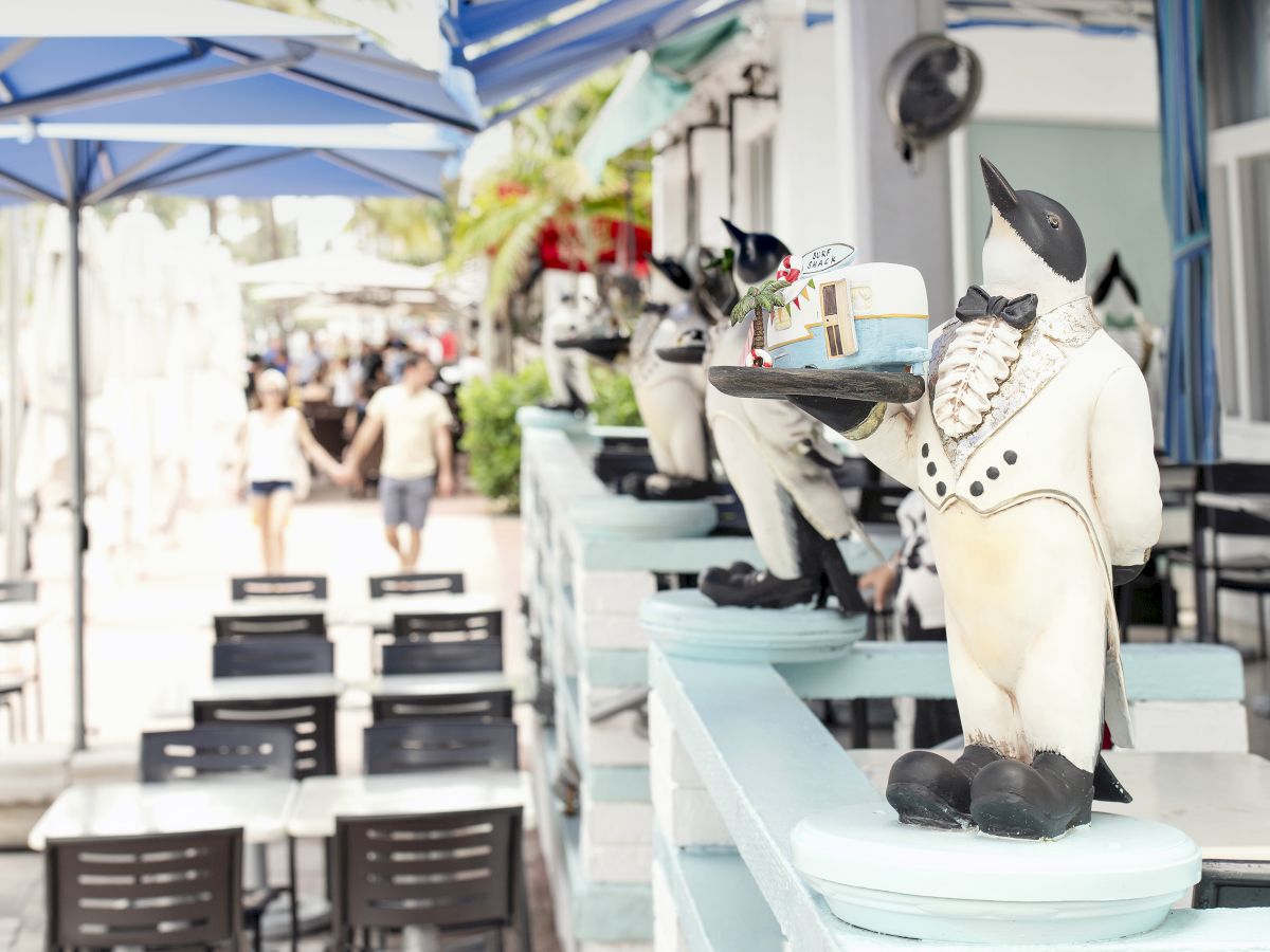 The image shows a patio area with tables and chairs, featuring penguin statues dressed as waiters holding trays, under outdoor umbrellas.