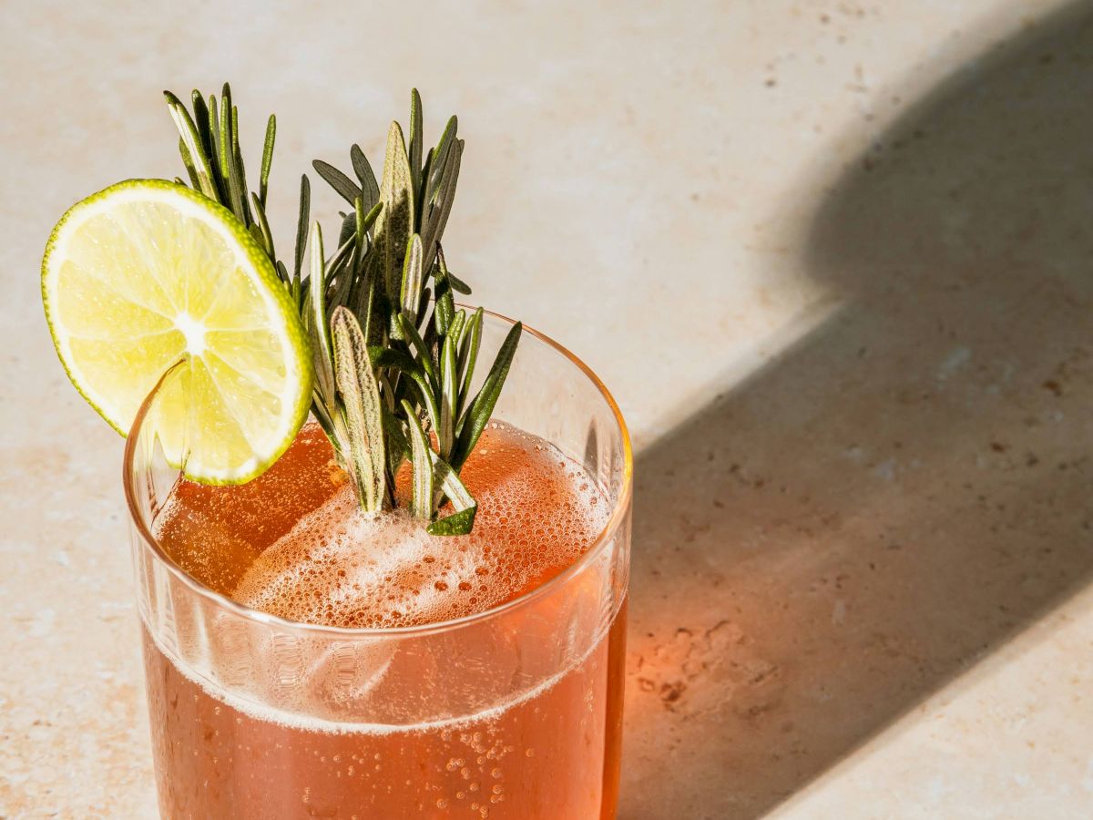 A cocktail in a glass with a lime slice and rosemary sprigs garnish sits on a light surface, casting a shadow.