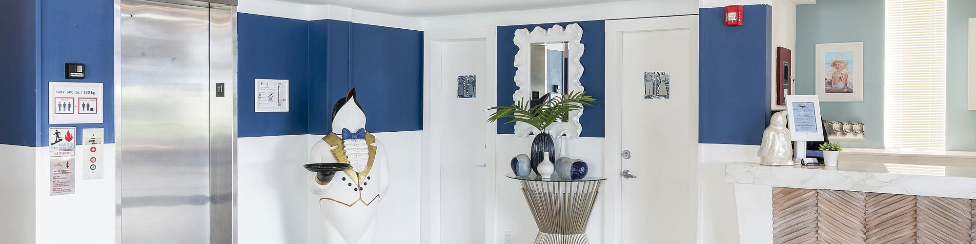 The image shows a hotel lobby with blue and white decor, an elevator, front desk, framed artwork, a decorative plant, and a modern chandelier.