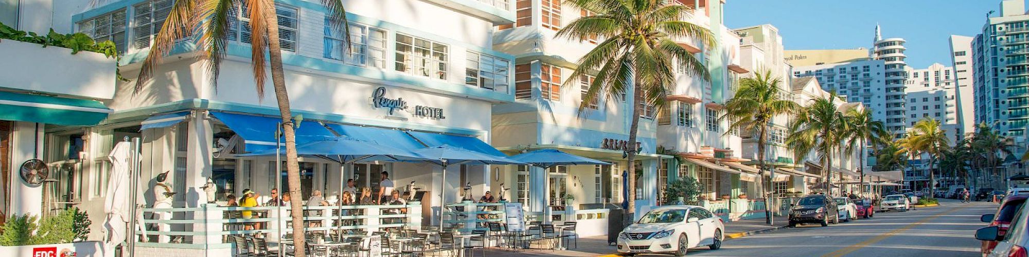 A sunny street with palm trees, art deco buildings, outdoor cafes, cars parked along the road, and people enjoying a leisurely day.