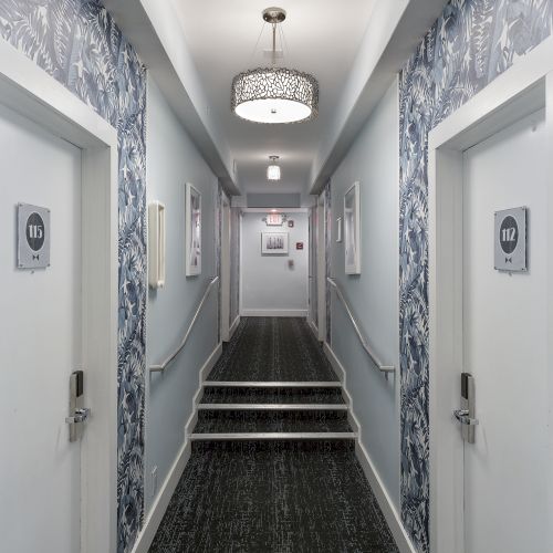 A hallway with tropical wallpaper, carpeted floor, two white doors labeled 112 and 113, and a light fixture on the ceiling, ending with a staircase.