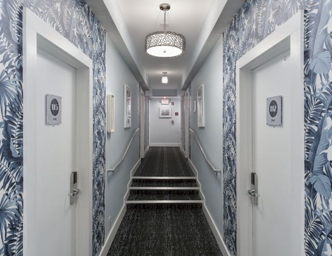 A hallway with tropical wallpaper, carpeted floor, two white doors labeled 112 and 113, and a light fixture on the ceiling, ending with a staircase.