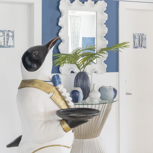 A penguin statue dressed as a butler holding a tray stands next to a table with vases and a plant, in front of two white doors and a decorative mirror.