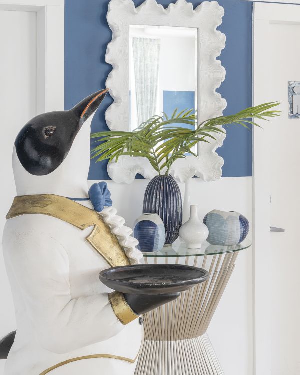 A penguin statue dressed as a butler holding a tray stands next to a table with vases and a plant, in front of two white doors and a decorative mirror.