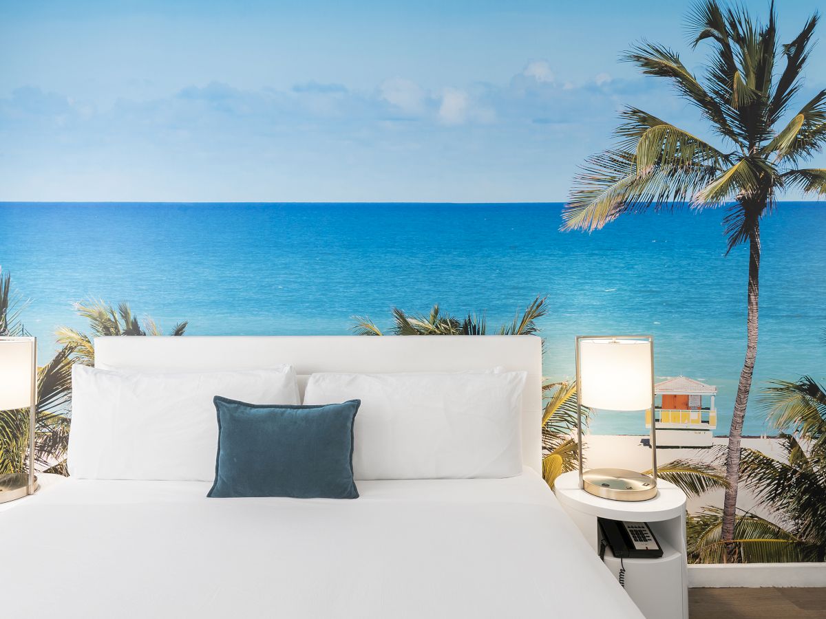 A bedroom with a beach-themed mural on the wall, depicting tropical palm trees and the ocean, complete with a neatly made bed and two bedside lamps.