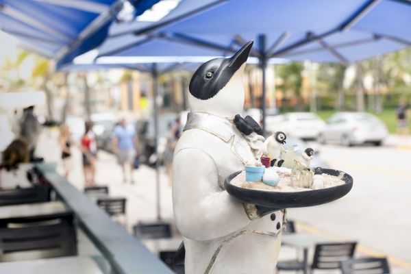 A penguin statue dressed as a waiter holding a tray with drinks stands outdoors under blue umbrellas, with people and cars in the background.