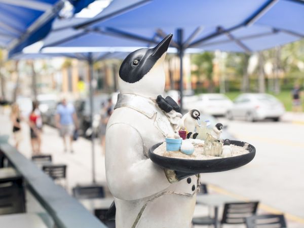 A penguin statue dressed as a waiter holding a tray with drinks stands outdoors under blue umbrellas, with people and cars in the background.