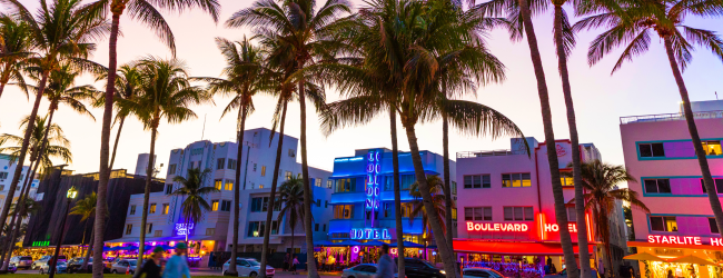 The image shows a lively evening scene with palm trees, colorful neon-lit buildings, and people walking, likely in a vibrant urban area near the coast.