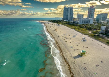 Aerial view of a beach with turquoise waters, sandy shores, people relaxing, and high-rise buildings in the background under a partly cloudy sky.