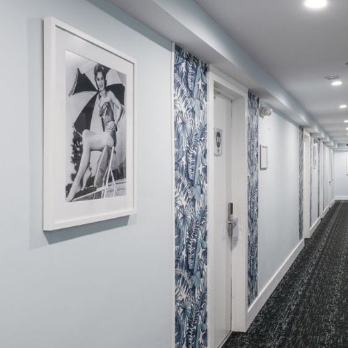 A hallway with patterned wallpaper, black-and-white framed photograph on the wall, blue carpeting, and doors on both sides ending at a distant exit.