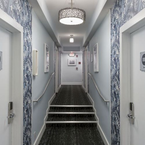 A hallway with blue floral wallpaper, stair steps, ceiling light fixture, two doors numbered 112 and 115, and framed pictures on the walls.