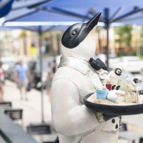 A statue of a penguin dressed as a waiter holding a tray with drinks, placed in an outdoor restaurant setting under blue umbrellas.