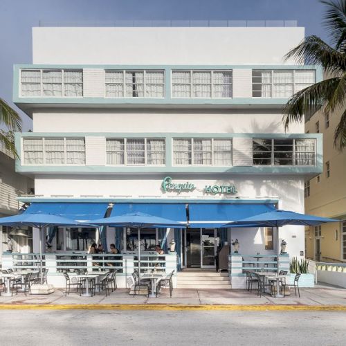 A white three-story building with a restaurant beneath blue awnings, palm trees, and outdoor seating on the sidewalk beneath a clear sky.