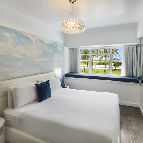 A modern bedroom with a white bed, blue accent pillow, bedside lamps, wall art, large window, mirrored wardrobe, and mounted flat-screen TV.